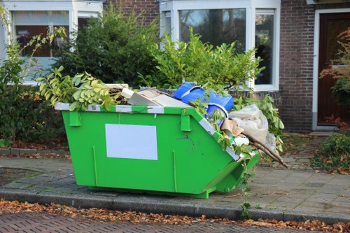 A large, green plastic skip overflowing with garden waste and miscellaneous rubbish, positioned on a paved driveway in front of a residential brick house with white-framed windows. The skip contains a mix of green leafy branches, small bushes, white cardboard boxes, blue plastic containers, and a white sack, all loosely piled and extending over the top edge. The surrounding environment includes a concrete curb at the base of the skip, fallen leaves scattered on the pavement, and shrubbery and plants growing along the house front, indicating a typical suburban setting likely in Camden Town. The scene is captured in natural daylight, emphasizing the textures of the garden debris and the smooth surface of the skip, which is part of a garden clearance service often provided by companies like Garden Clearance Camden Town for local residents needing rubbish removal from gardens or outdoor spaces in the area.