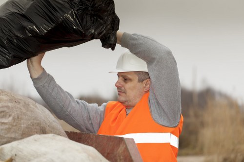 A male waste collection worker with light skin and short dark hair, wearing a white helmet and an orange high-visibility vest over a grey long-sleeved shirt, is lifting a large black rubbish bag above his head. He is positioned outdoors in an open environment with blurred natural background, possibly indicating a rural or suburban area. The worker appears focused on his task of collecting or removing waste materials. In the foreground, there are large, light-colored bags and waste containers, suggesting a rubbish removal activity. The overall scene depicts a professional waste management operation, aligning with services offered by companies such as Garden Clearance Camden Town, specialising in rubbish disposal and clearance tasks in north London.