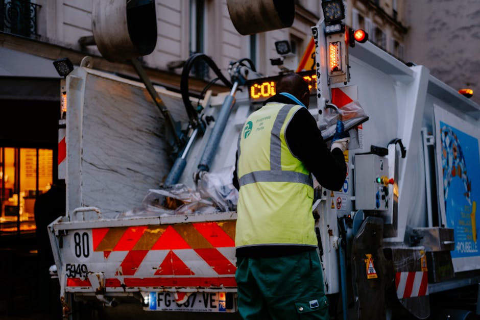 A person wearing a dark grey cap, black face mask, and an orange high-visibility jacket with green reflective stripes stands on a city sidewalk, sorting through a collection of clear and black rubbish
